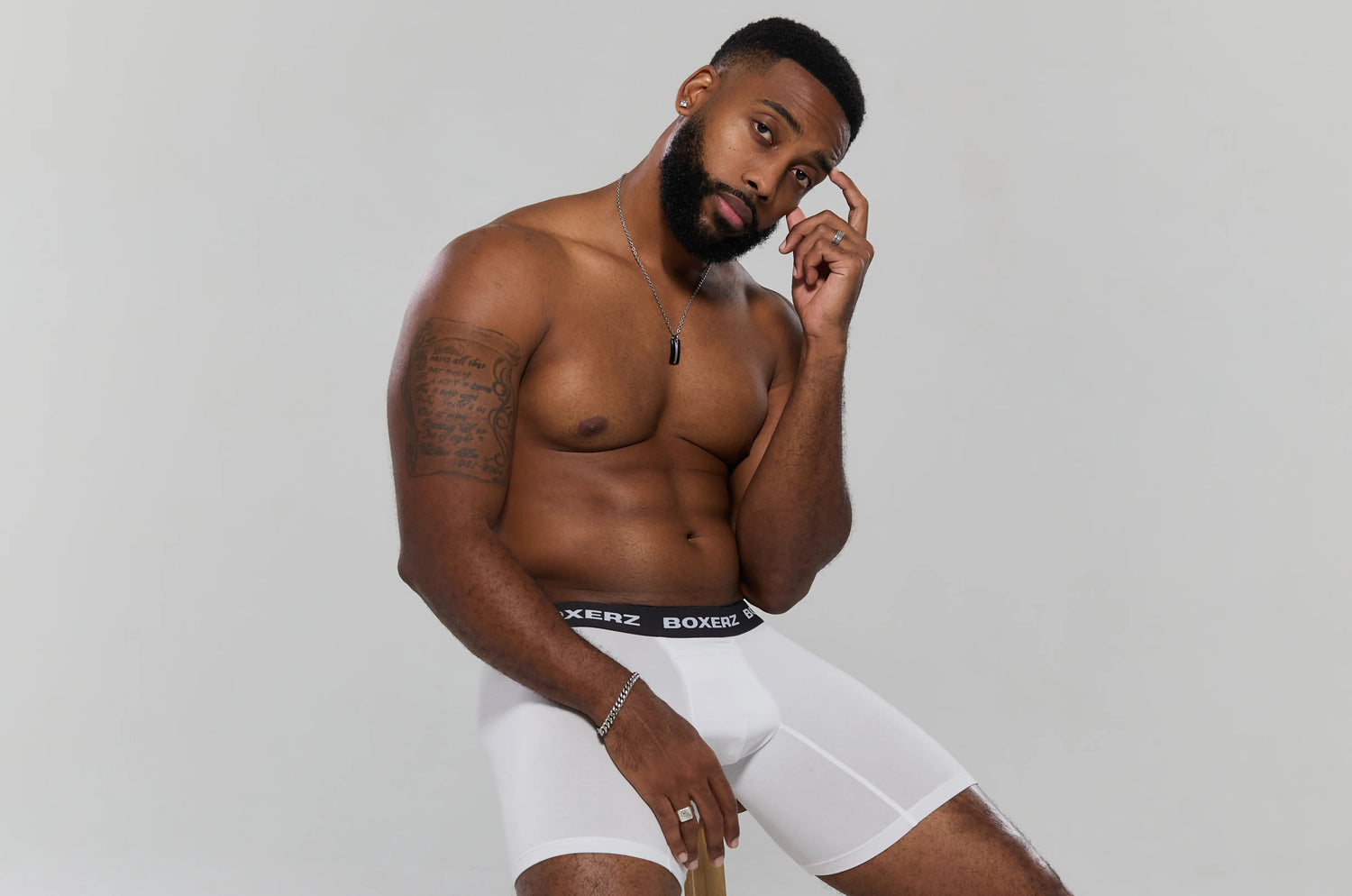 A confident male model poses shirtless in white BOXERZ boxer briefs, seated against a light grey studio backdrop.